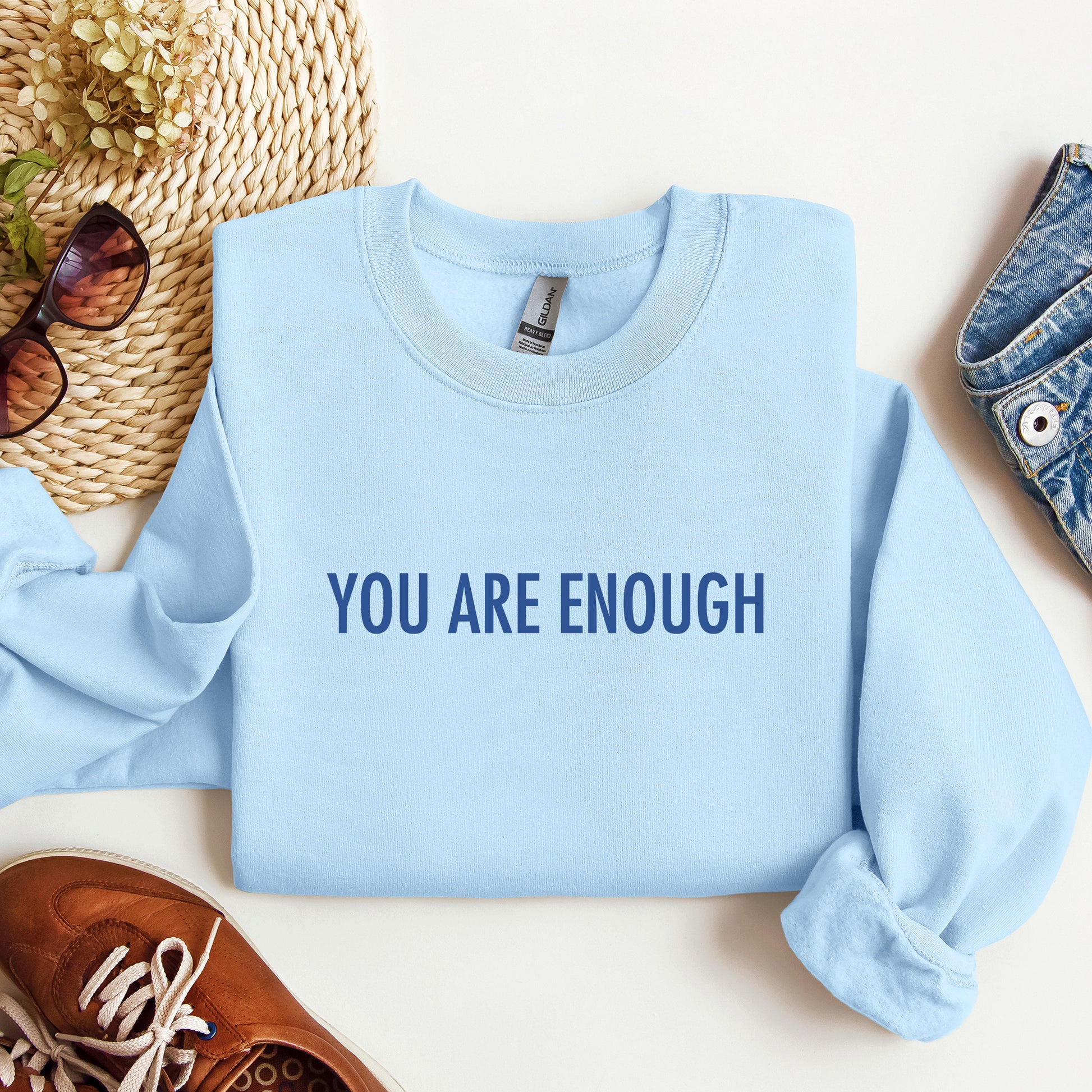 A light blue crewneck sweatshirt with the text 'YOU ARE ENOUGH' printed in navy blue across the chest, styled in a flat lay with jeans, sunglasses, a woven hat, and brown shoes.