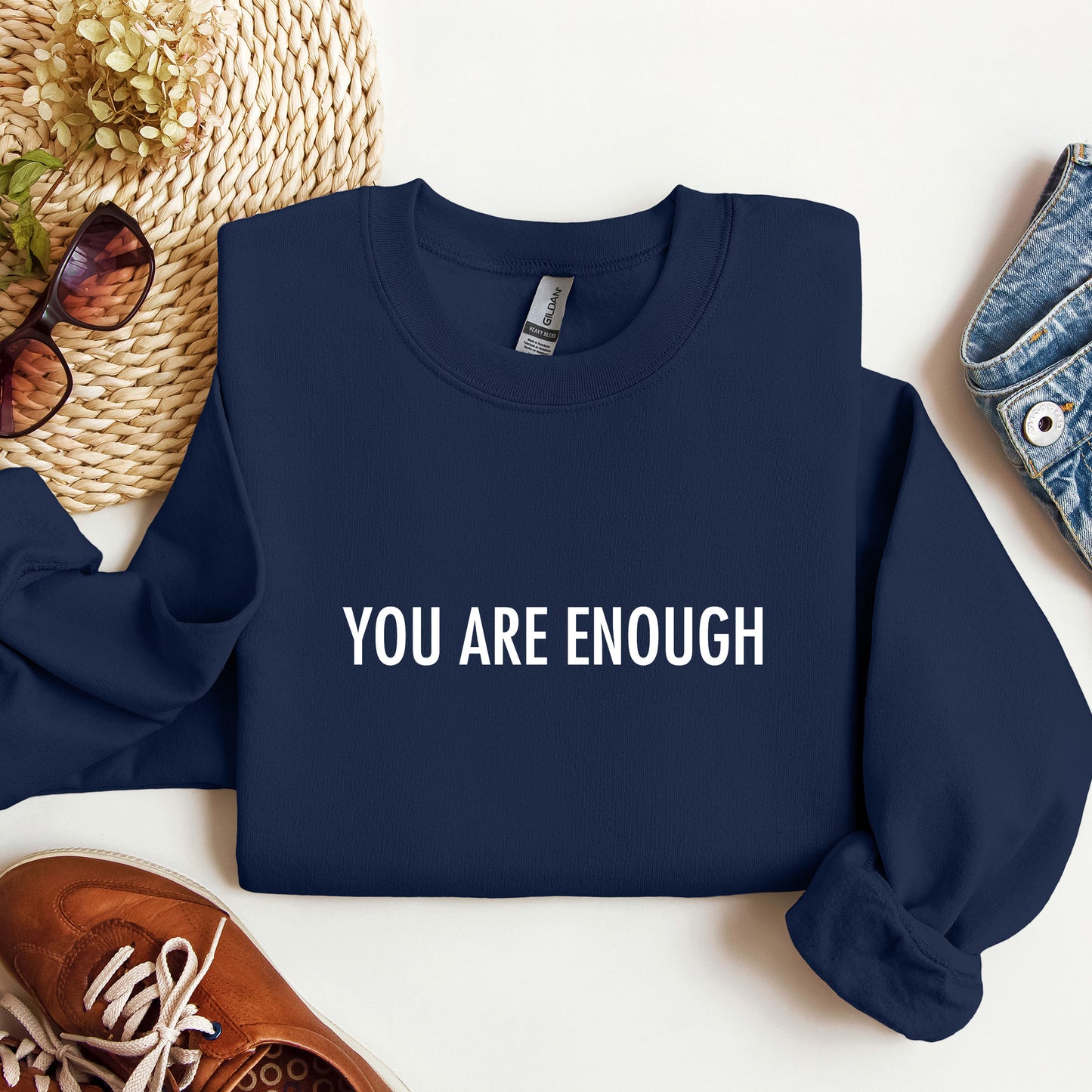 A navy blue crewneck sweatshirt with the phrase 'YOU ARE ENOUGH' printed in white capital letters across the chest. The sweatshirt is presented in a styled flat-lay arrangement on a white surface, surrounded by a woven straw hat, sunglasses, brown leather shoes, and blue denim.