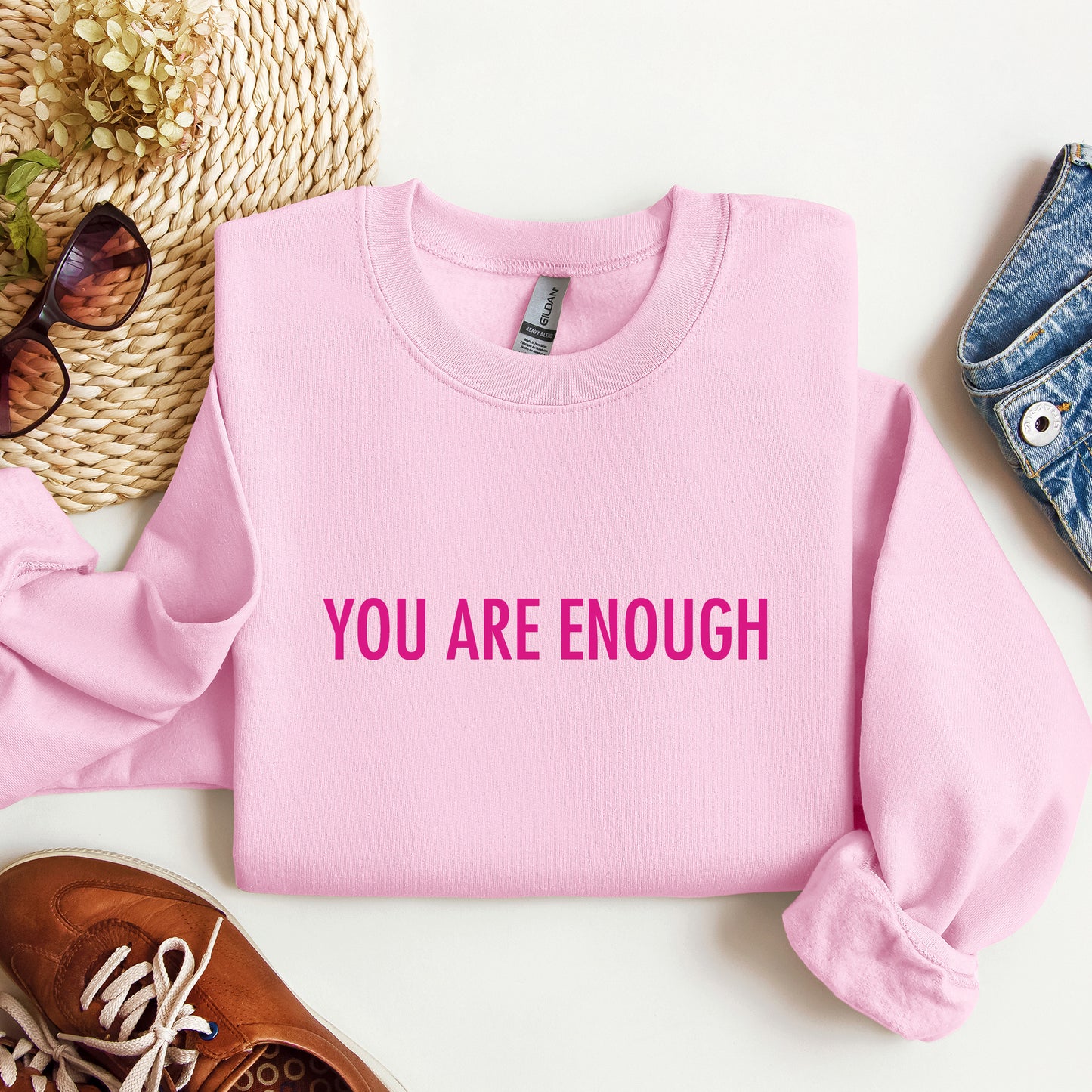 A folded light pink crewneck sweatshirt with the words 'YOU ARE ENOUGH' printed in bright pink across the chest, styled in a flat lay with a straw hat, sunglasses, a brown shoe, and blue jeans on a white background.