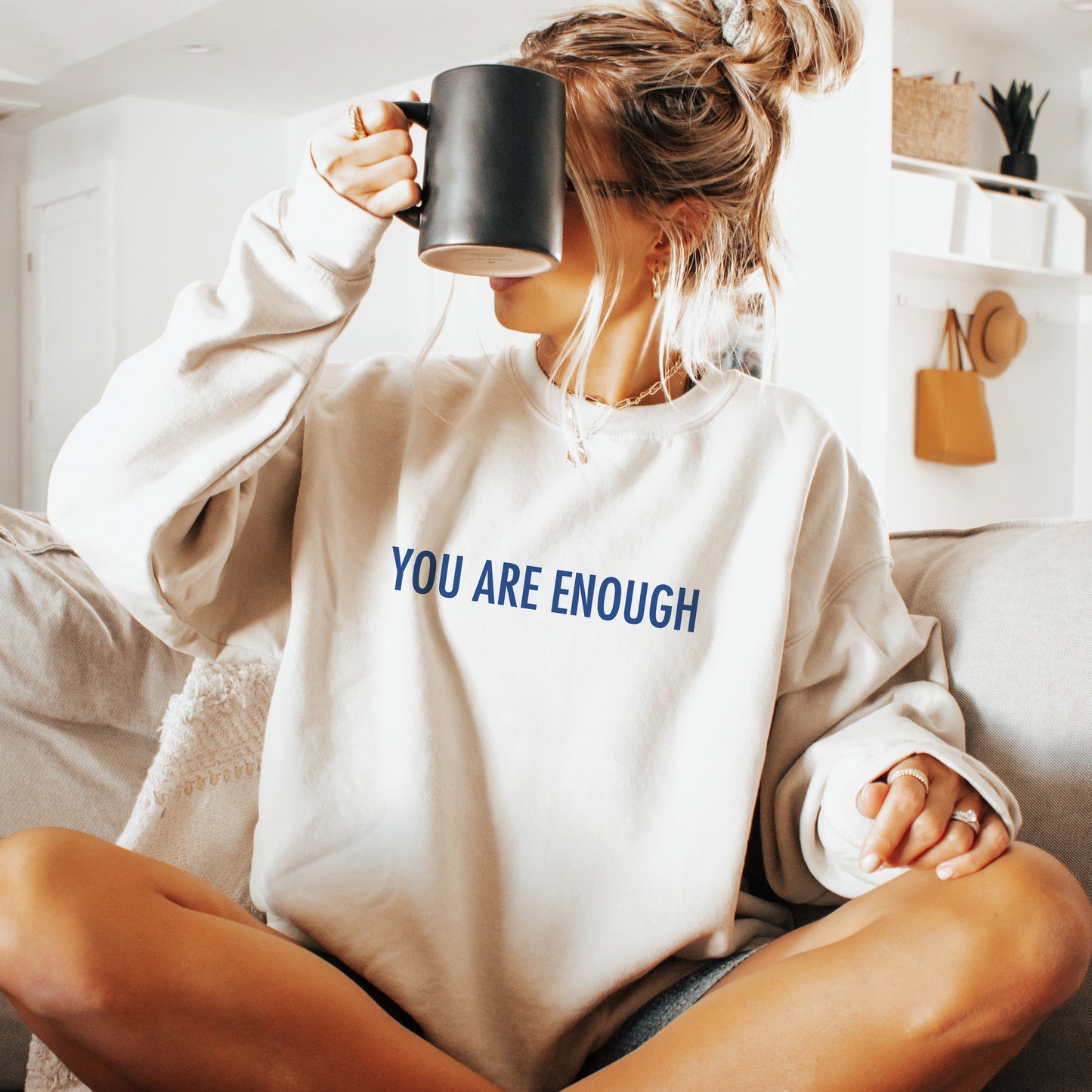 A woman sitting on a couch in a bright living room, wearing an oversized cream sweatshirt with the words 'YOU ARE ENOUGH' printed in blue across the chest, holding a black mug to her face.