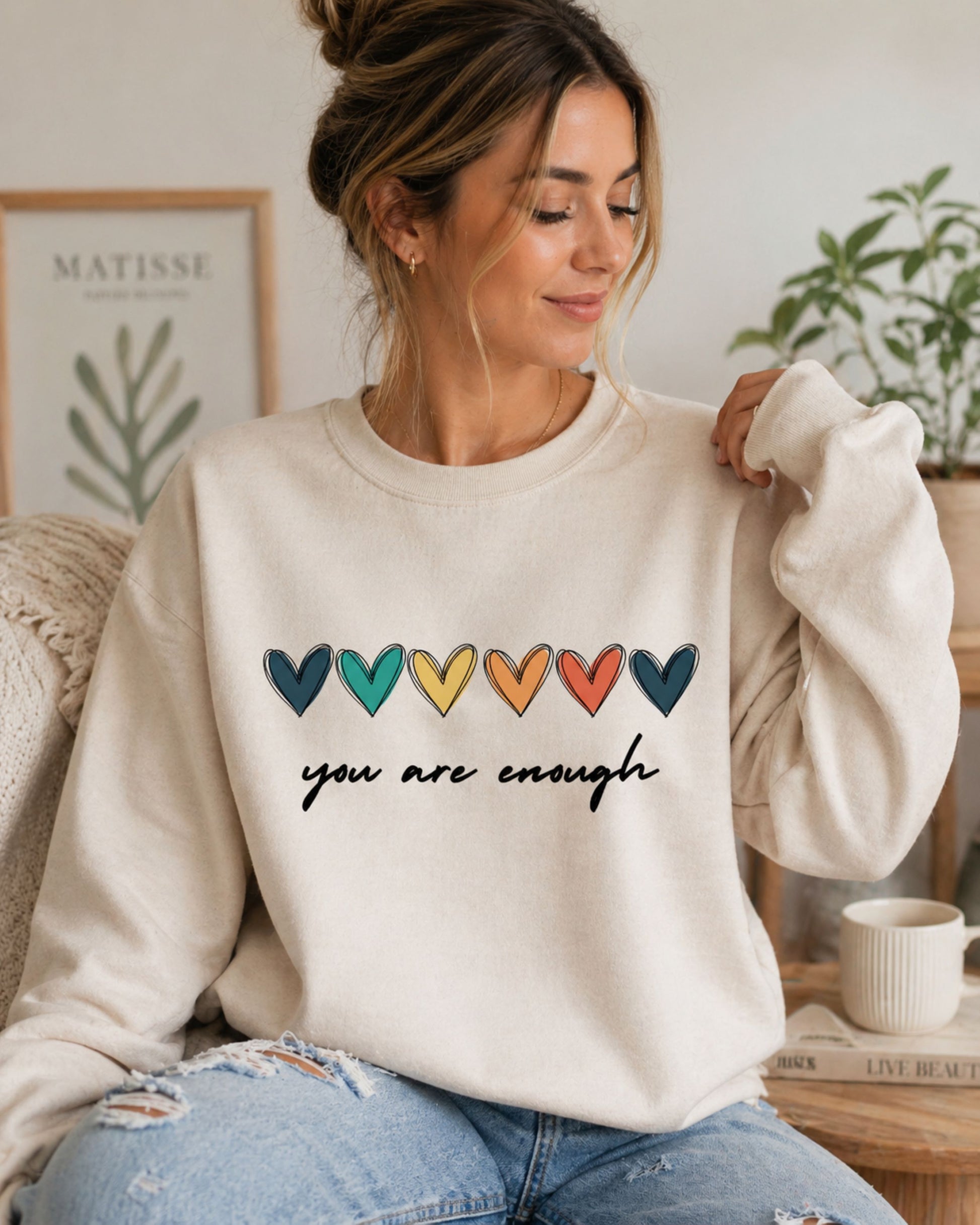 Woman wearing a beige sweatshirt with colorful heart design and 'you are enough' text, sitting in a cozy room.
