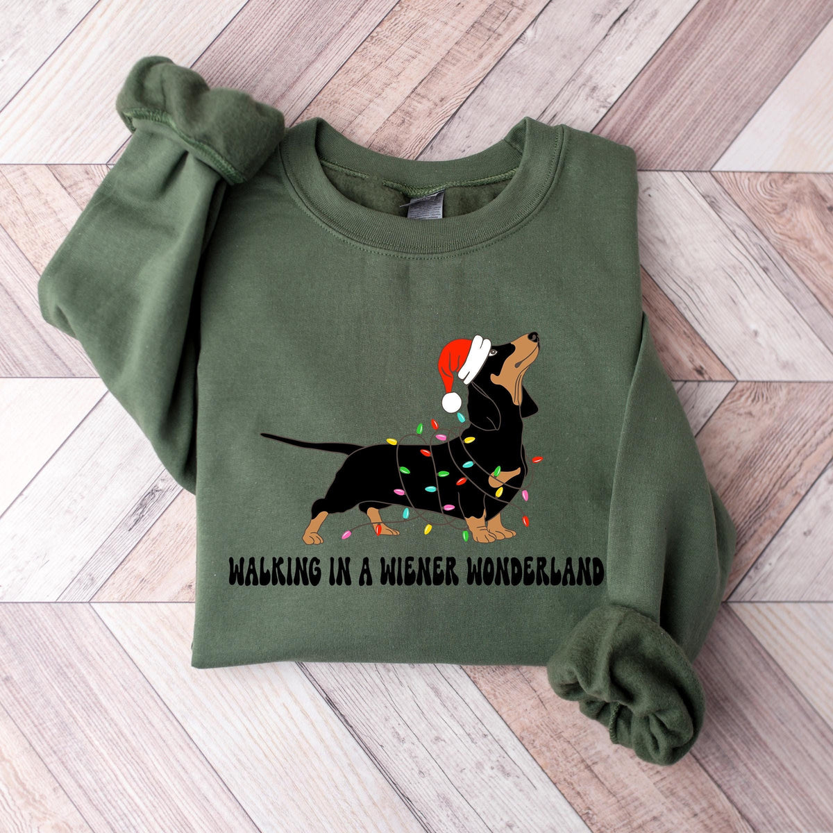 A military green Gildan sweatshirt with a picture of a dachshund tangled in christmas lights and wearing a santa hat.  Beneath the picture are the words, Walking in a Wiener Wonderland