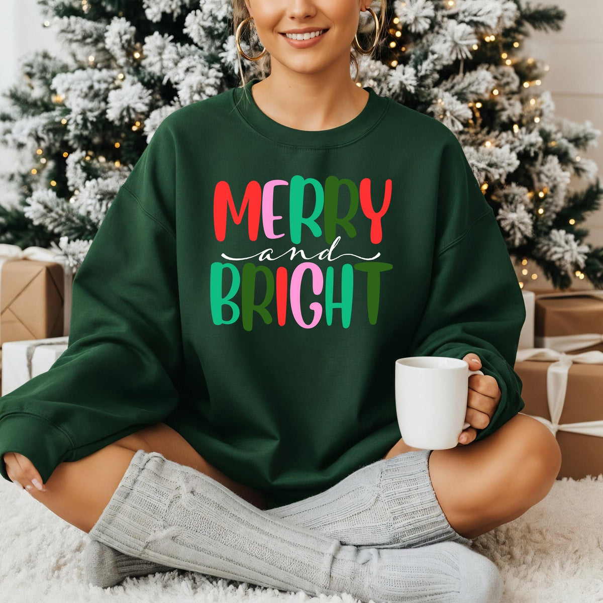 A person sits in front of a lit Christmas tree wearing a dark green sweatshirt with colorful Merry and Bright text.
