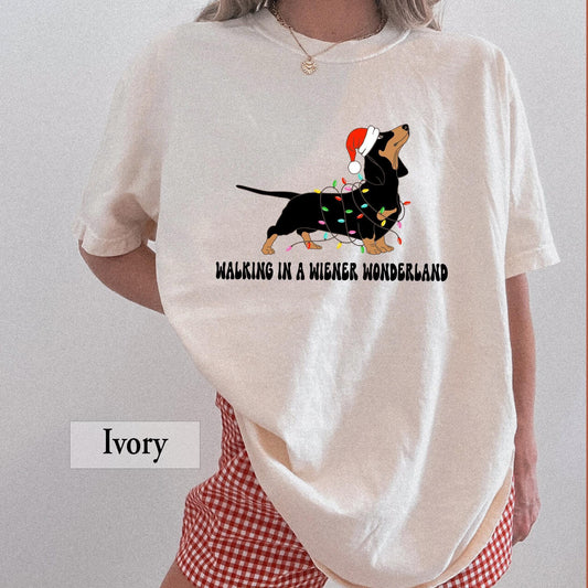 An ivory Comfort Colors shirt with a picture of a dachshund on it.  The dog is tangled in christmas lights and is wearing a santa hat.  Below the picture it says Walking in a Wiener Wonderland.