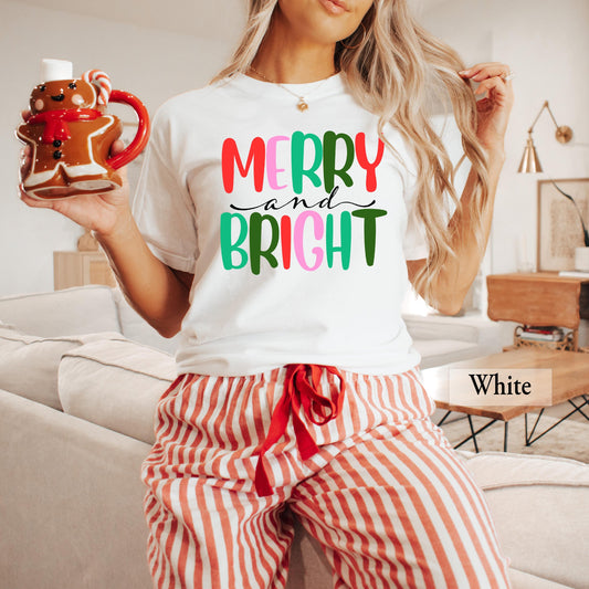 A person wearing a white t-shirt with colorful Merry and Bright text, paired with red and white striped pajama pants, holding a gingerbread mug.