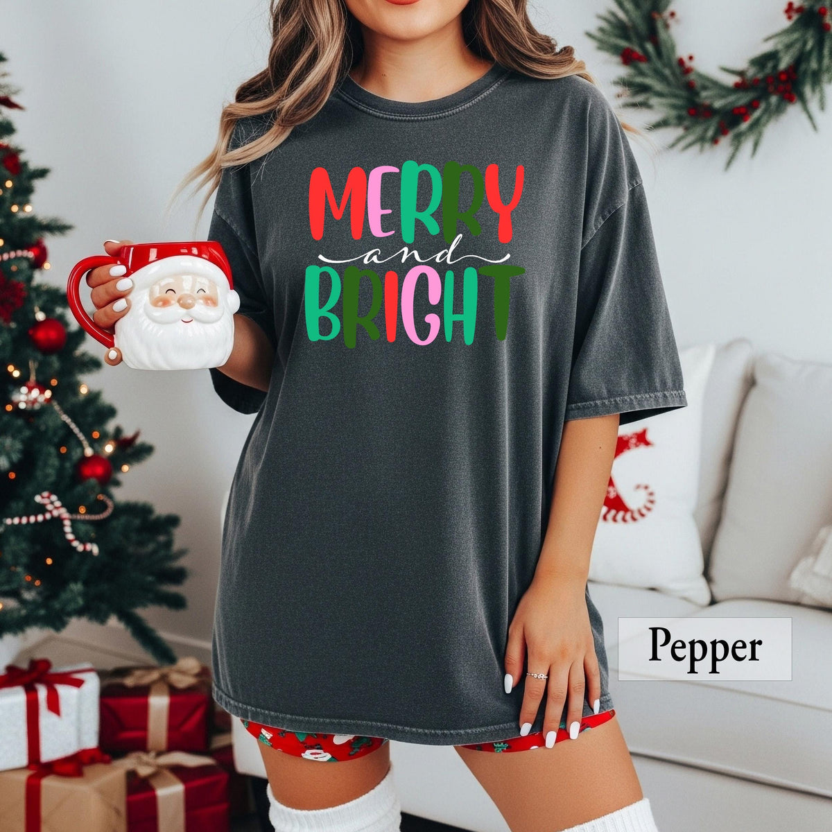 A person wearing a pepper grey t-shirt with colorful Merry and Bright text, sitting on a white couch next to a Santa mug.