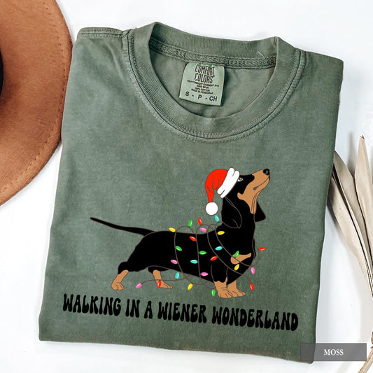 A moss Comfort Colors shirt with a picture of a dachshund on it.  The dog is tangled in christmas lights and is wearing a santa hat.  Below the picture it says Walking in a Wiener Wonderland.