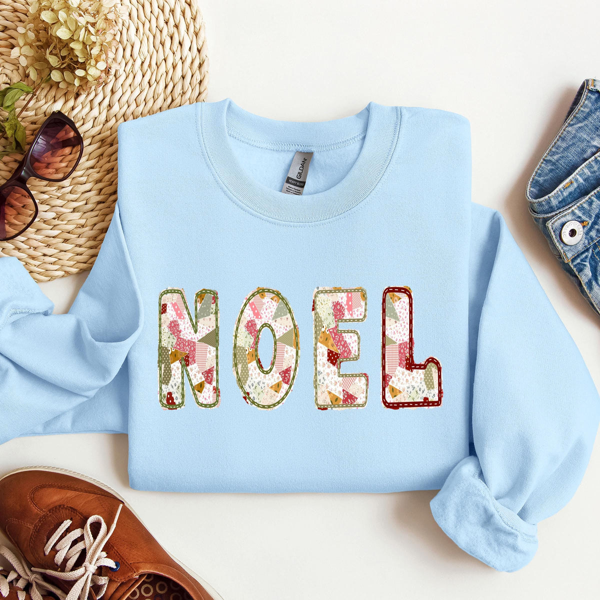 A light blue Gildan crewneck sweatshirt with NOEL printed on it in a muted sage, maroon, and white patchwork pattern.