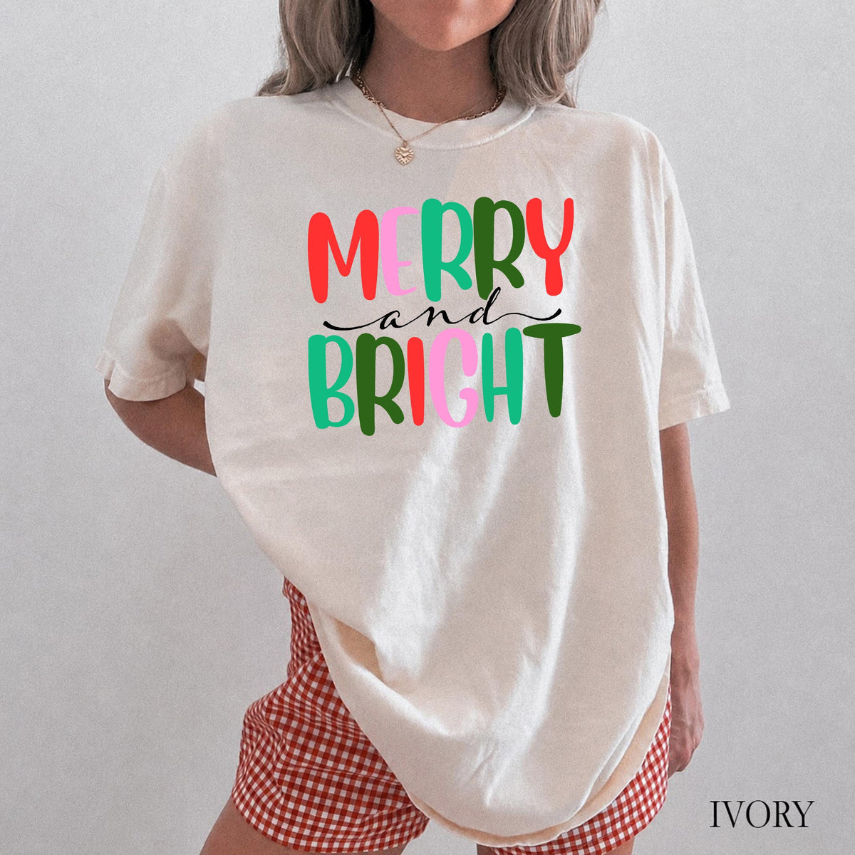 A person wearing an ivory t-shirt with colorful Merry and Bright text and red-and-white checkered shorts.