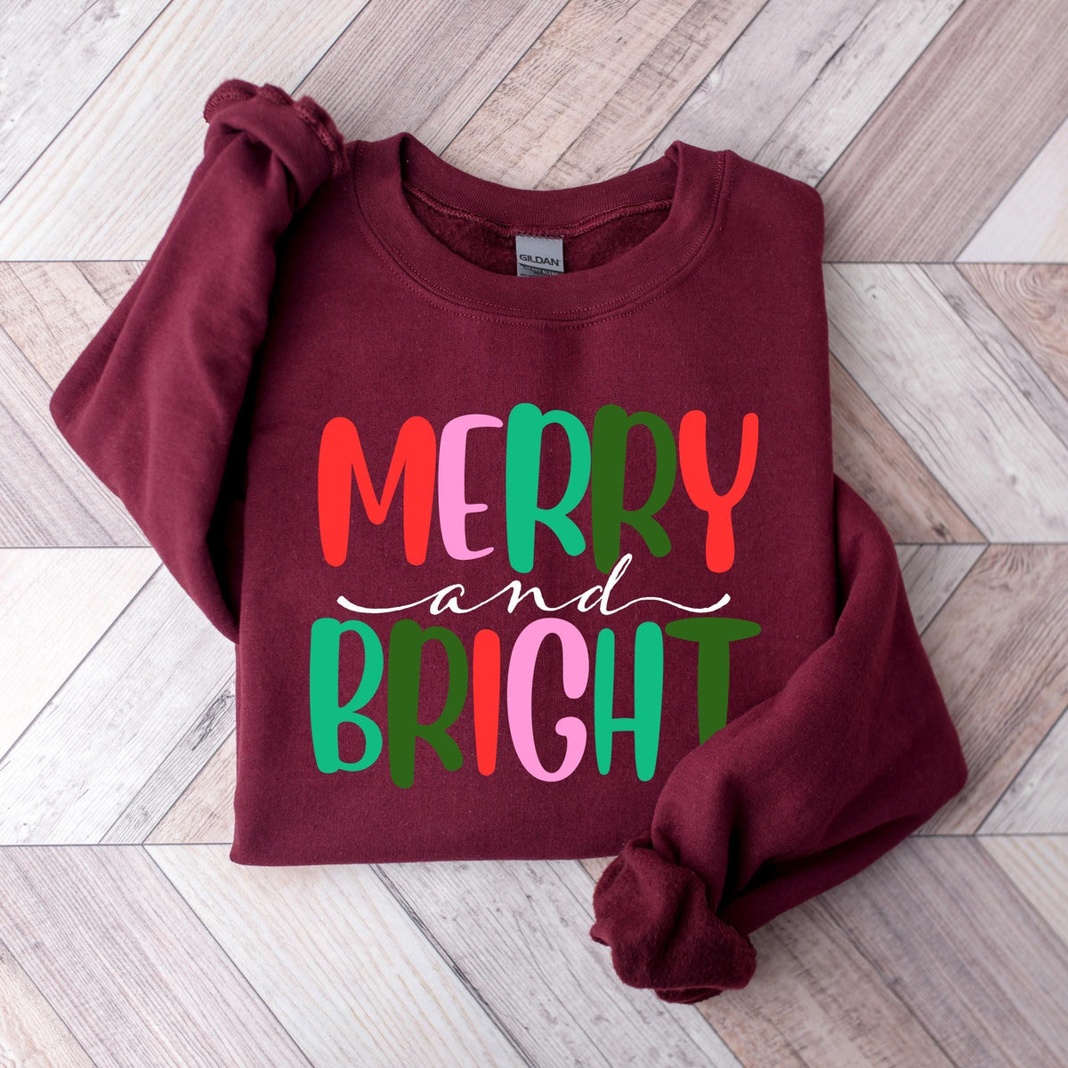 A maroon crewneck sweatshirt with the text Merry and Bright laid flat on a wooden surface.