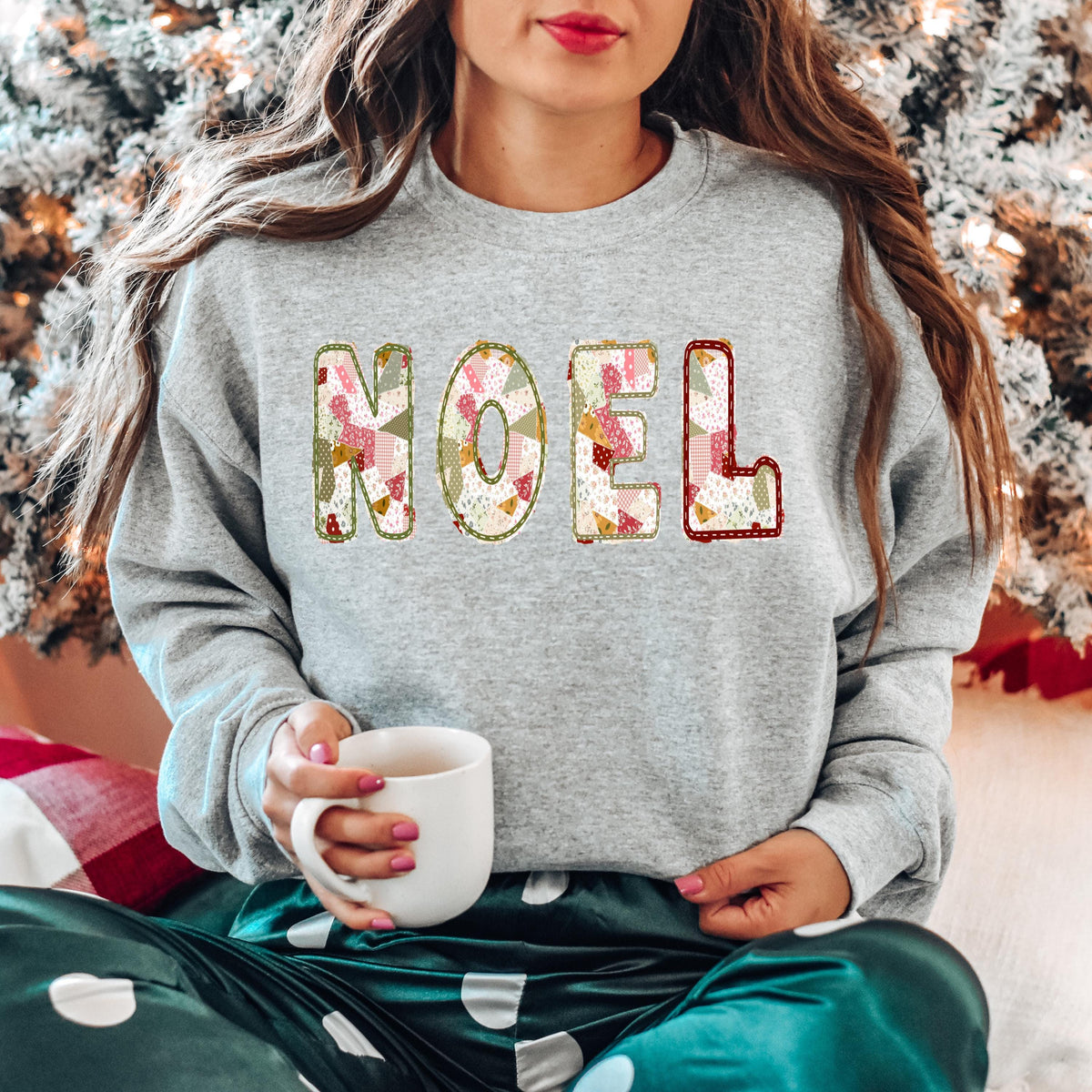 A woman wearing a heather grey Gildan crewneck sweatshirt with NOEL printed on it in a muted sage, maroon, and white patchwork pattern.